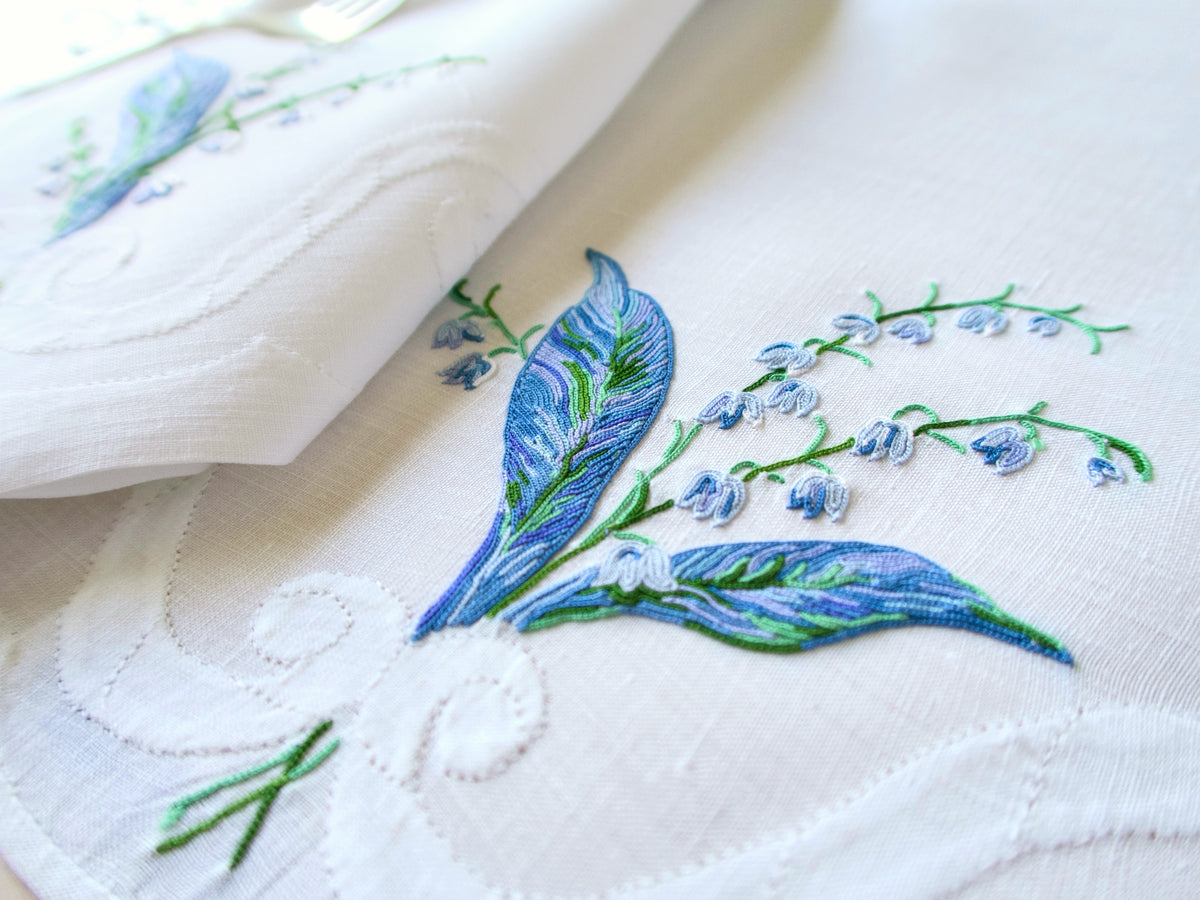 Close-up of D. Porthault Lily of the Valley Point de Beauvais embroidery showing layered blue and green thread detail on white linen placemat.