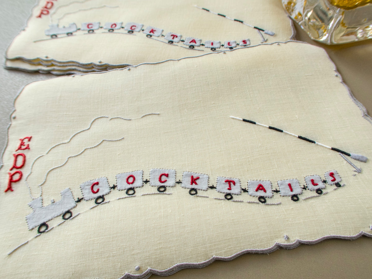 Two vintage Madeira cocktail napkins with a hand embroidered and appliquéd steam train whose cars spell COCKTAILS beside a striped crossing gate