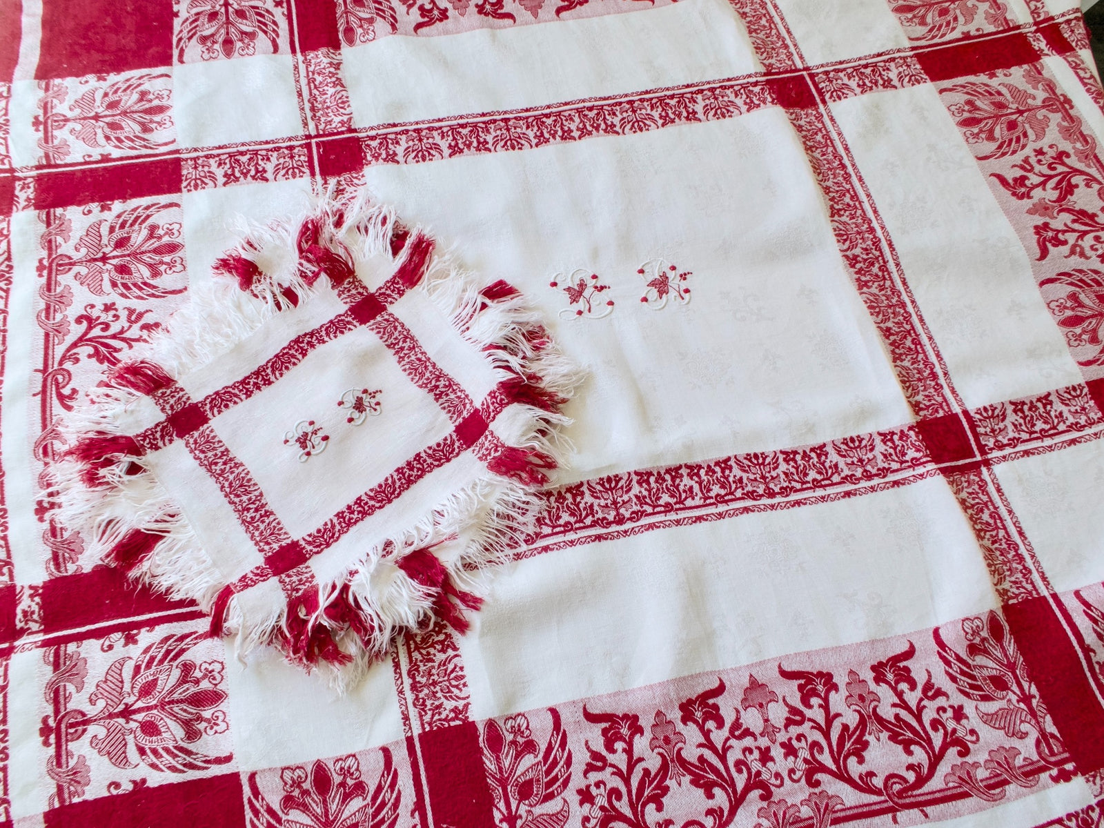 French red and white damask tablecloth with a matching fringed napkin, showing woven floral borders and embroidered LB monograms.