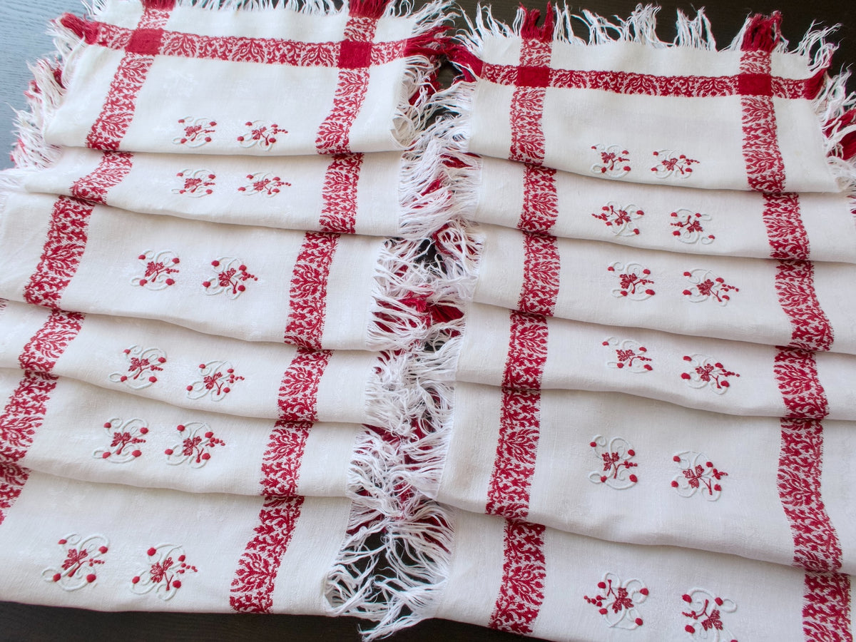 Twelve matching fringed napkins arranged in two rows, showing their woven red borders and embroidered monograms.