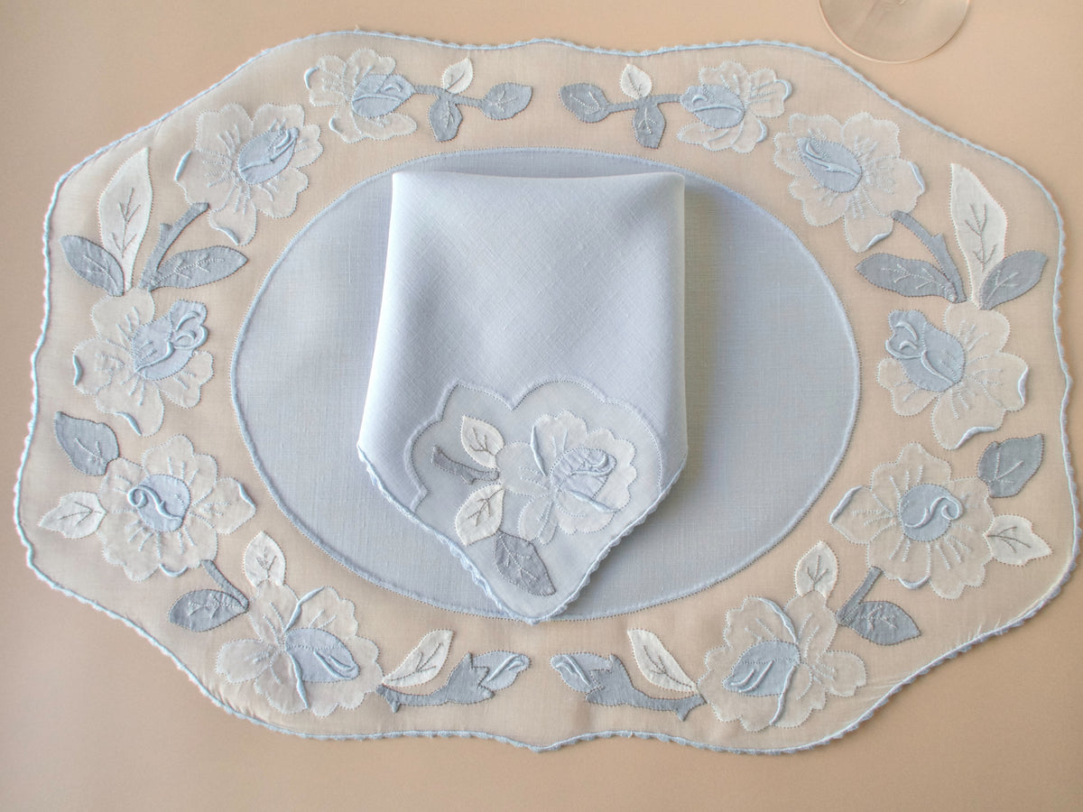 Vintage Madeira pale blue organdy placemat set for 8 showing complete oval shape with appliqué rose border, folded napkin with matching motif, beige background.