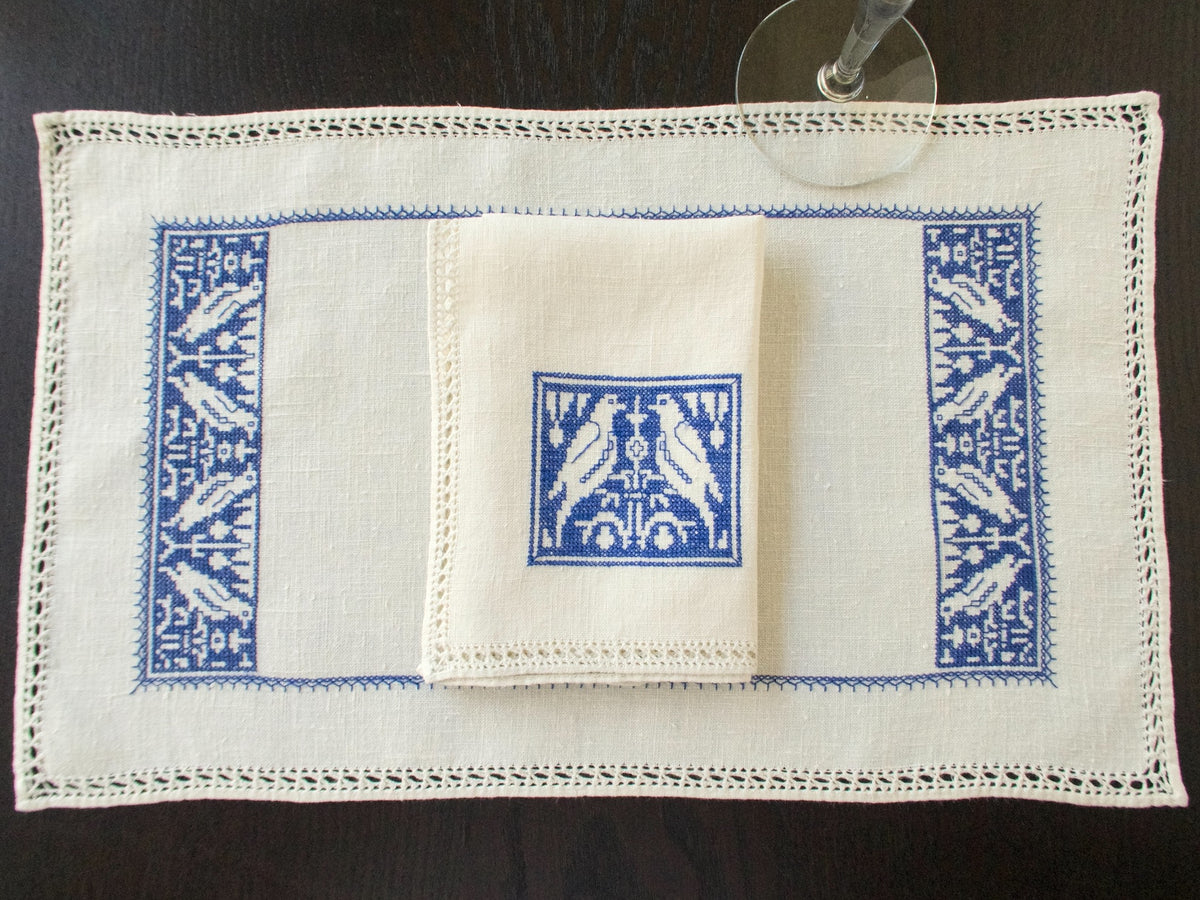 Overhead view of blue Assisi embroidery on a natural linen placemat, with matching napkins and drawn-thread edges.