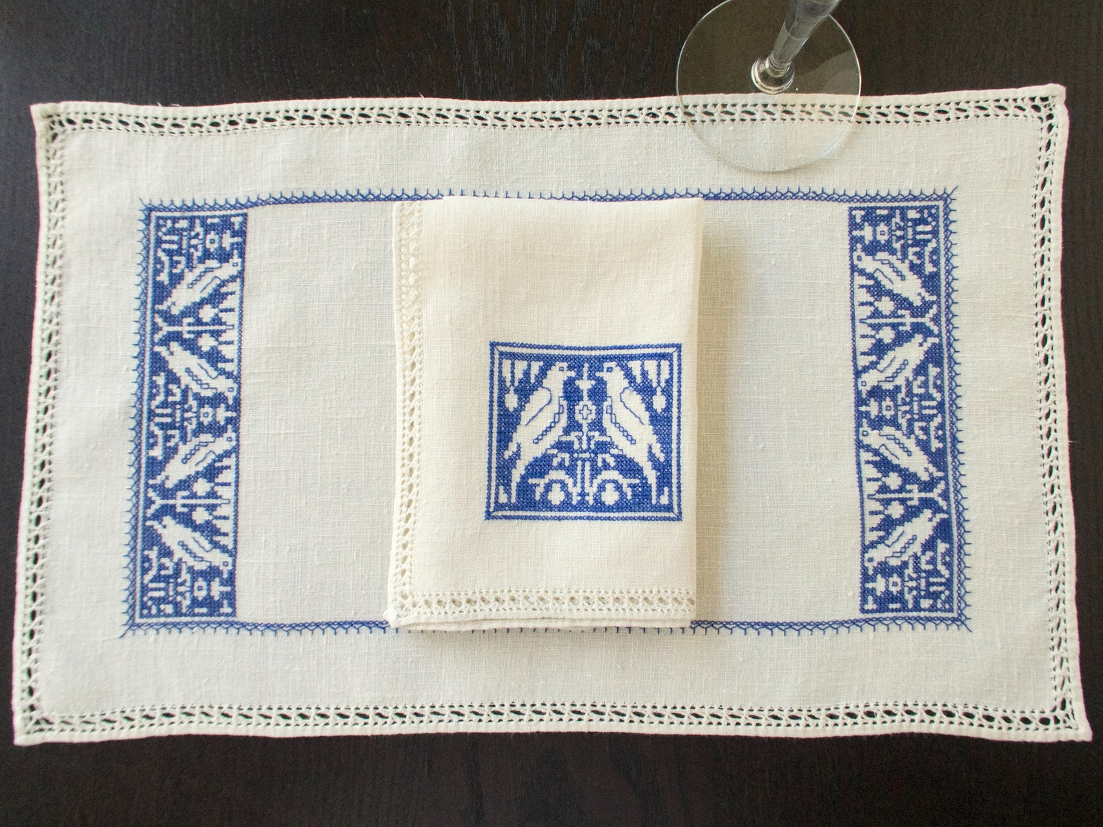 Overhead view of blue Assisi embroidery on a natural linen placemat, with matching napkins and drawn-thread edges.