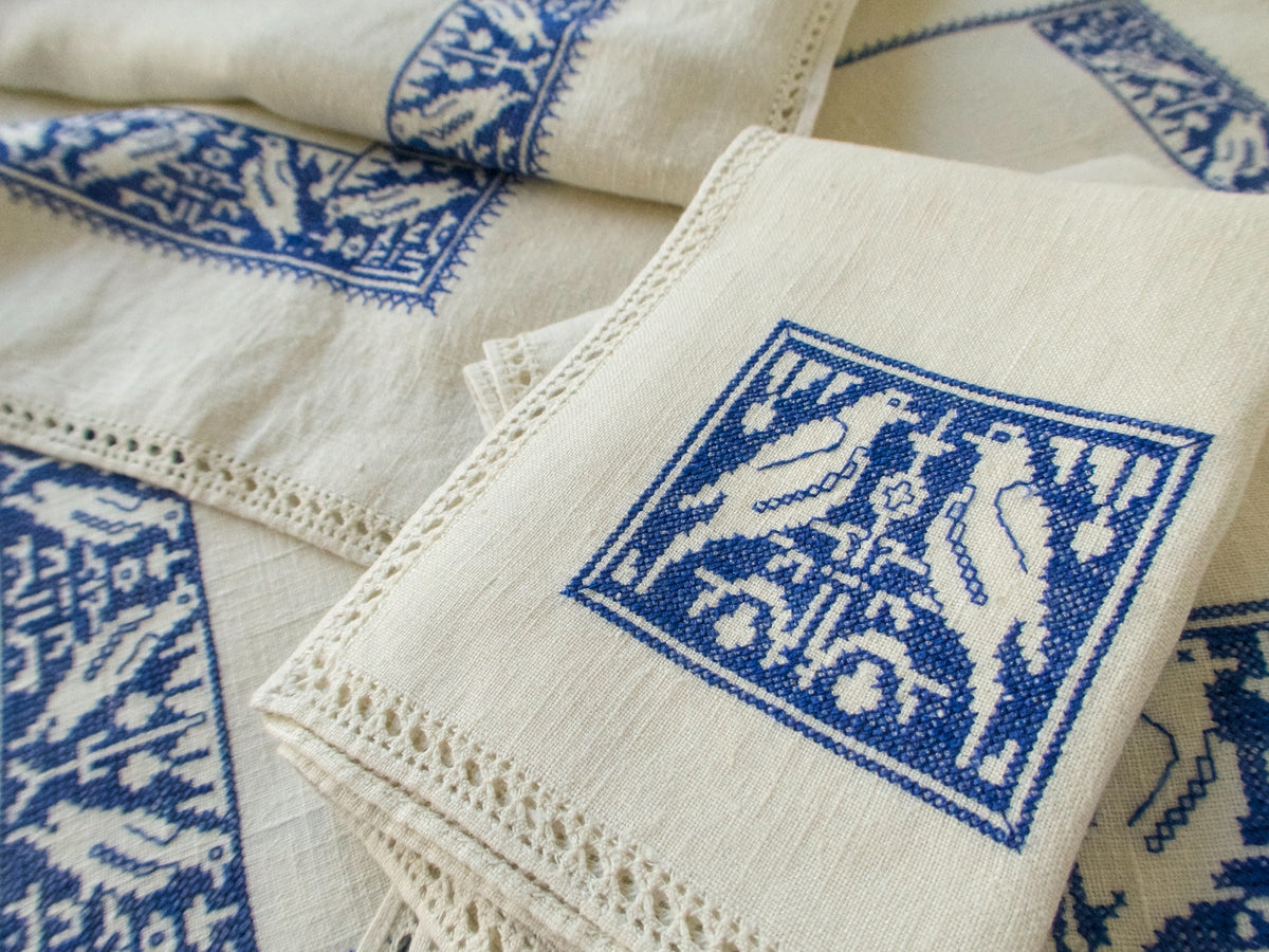 Close-up of a linen napkin with blue Assisi embroidery depicting two birds and a drawn-thread hemstitched edge.