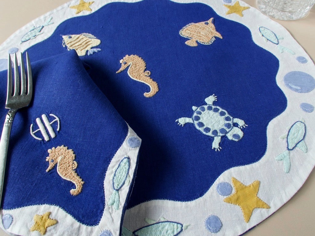 View of hand embroidered sea creatures on vintage Léron deep blue linen placemat, with seahorse and starfish detail, with folded napkin.