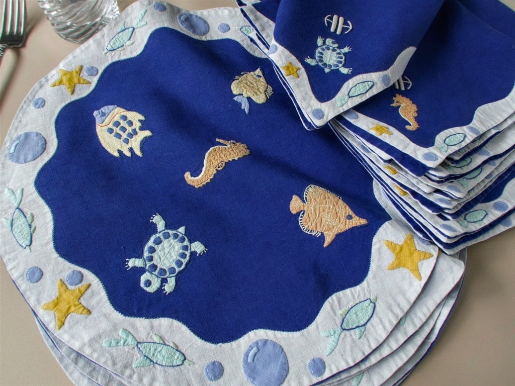 Vintage Léron deep blue linen placemats stacked and fanned, showing hand embroidered sea creatures and scalloped border.