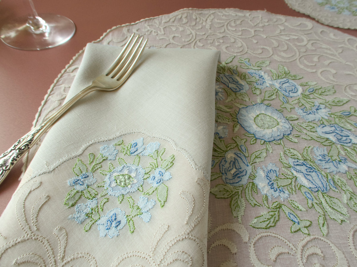 Close up of folded linen napkin and Madeira organdy placemat, blue flower and scrolling motif shadow work embroidery