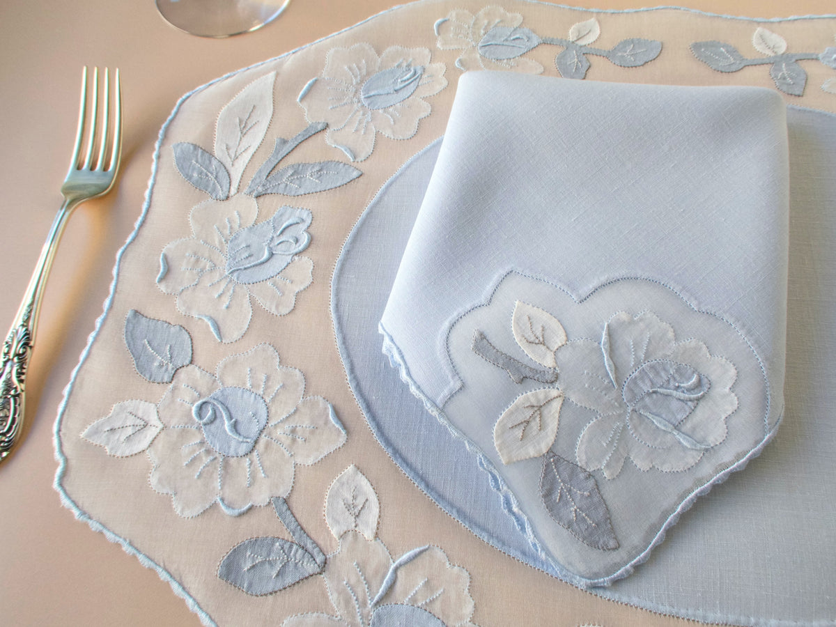 Detail of vintage Madeira pale blue organdy placemat showing large two-tone appliqué rose and leaf motifs with scalloped edge, Irish linen napkin alongside.