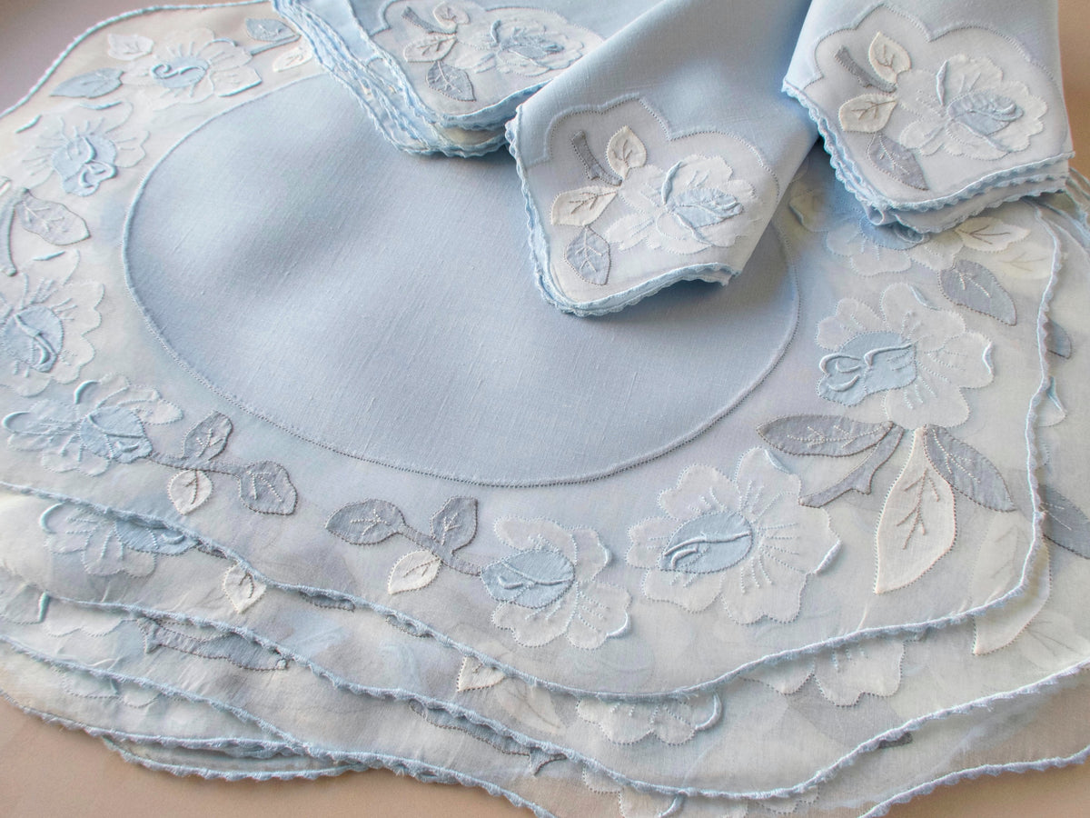 Stack of vintage Madeira pale blue organdy placemats with white and blue appliqué rose border, folded napkins showing set of 8, beige background.​​​​​​​​​​​​​​​​