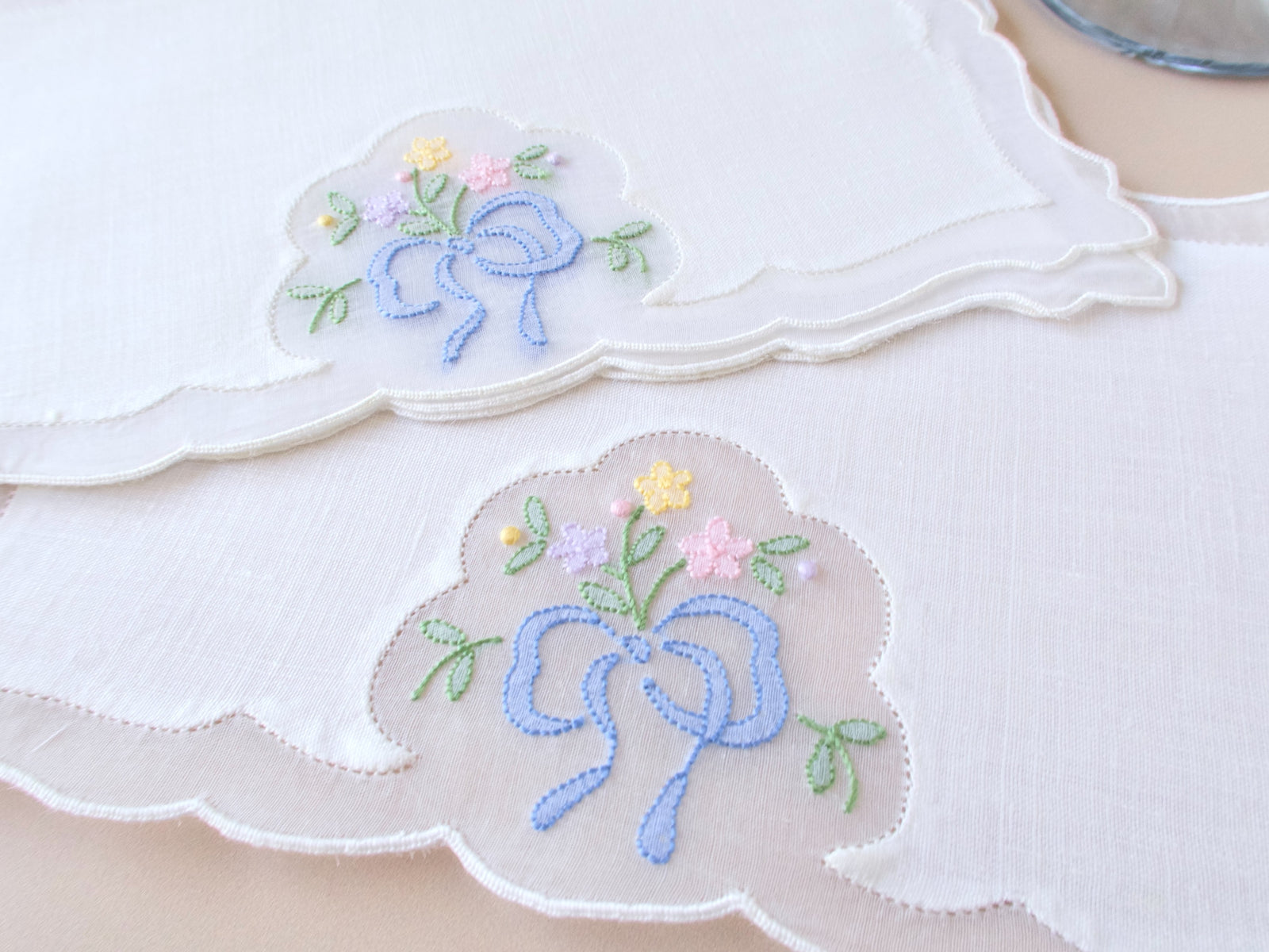 Close view of vintage Madeira cocktail napkin with blue ribbon bow and posy embroidery in yellow and pink on white linen and organdy, scalloped border detail.​​​​​​​​​​​​​​​​
