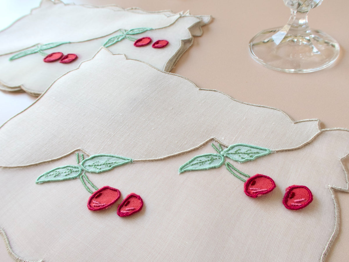 Close view of vintage Madeira trembler cocktail napkin showing three-dimensional detached red cherry embroidery with green leaves on white linen and organdy, scalloped edge detail visible.​​​​​​​​​​​​​​​​