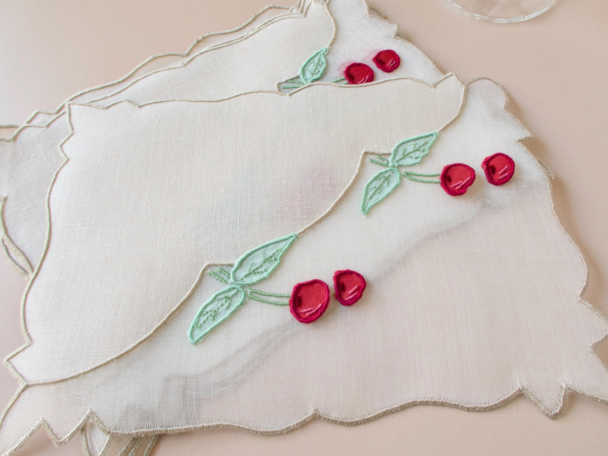 Set of six vintage Madeira cocktail napkins in white linen and organdy, hand embroidered with detached trembler cherries and green leaves in three-dimensional relief, fanned on a neutral surface with a crystal cocktail glass.