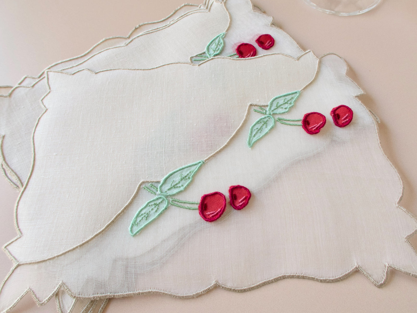 Set of six vintage Madeira cocktail napkins in white linen and organdy, hand embroidered with detached trembler cherries and green leaves in three-dimensional relief, fanned on a neutral surface with a crystal cocktail glass.