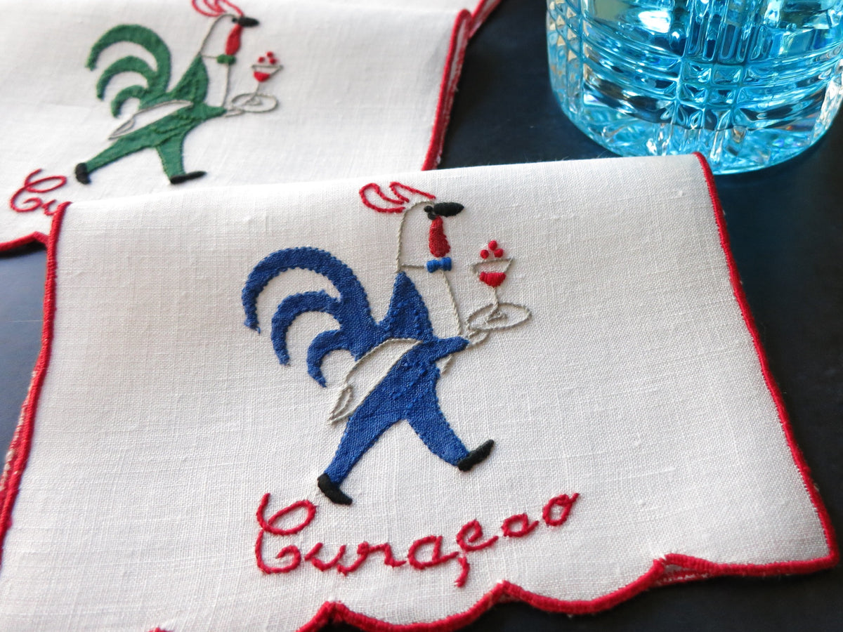Vintage Madewira embroidered rooster cocktail napkin, white linen with red trim, placed on a table.