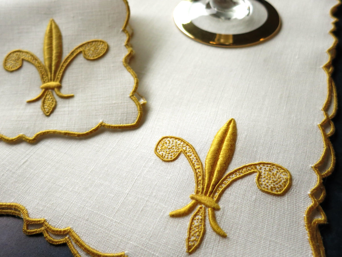 Close-up of a vintage linen cocktail napkin with raised gold fleur-de-lis embroidery and scalloped edges, hand embroidered in Madeira.