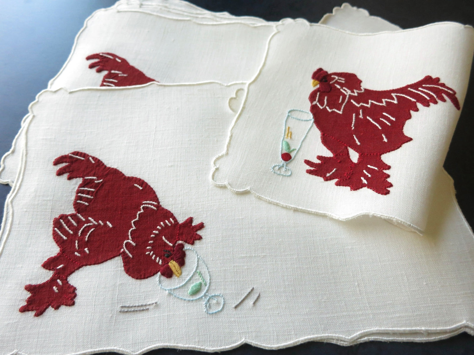 Stacked vintage Madeira cocktail napkins with red appliqué roosters and garnishes showing scalloped border on linen​​​​​​​​​​​​​​​​