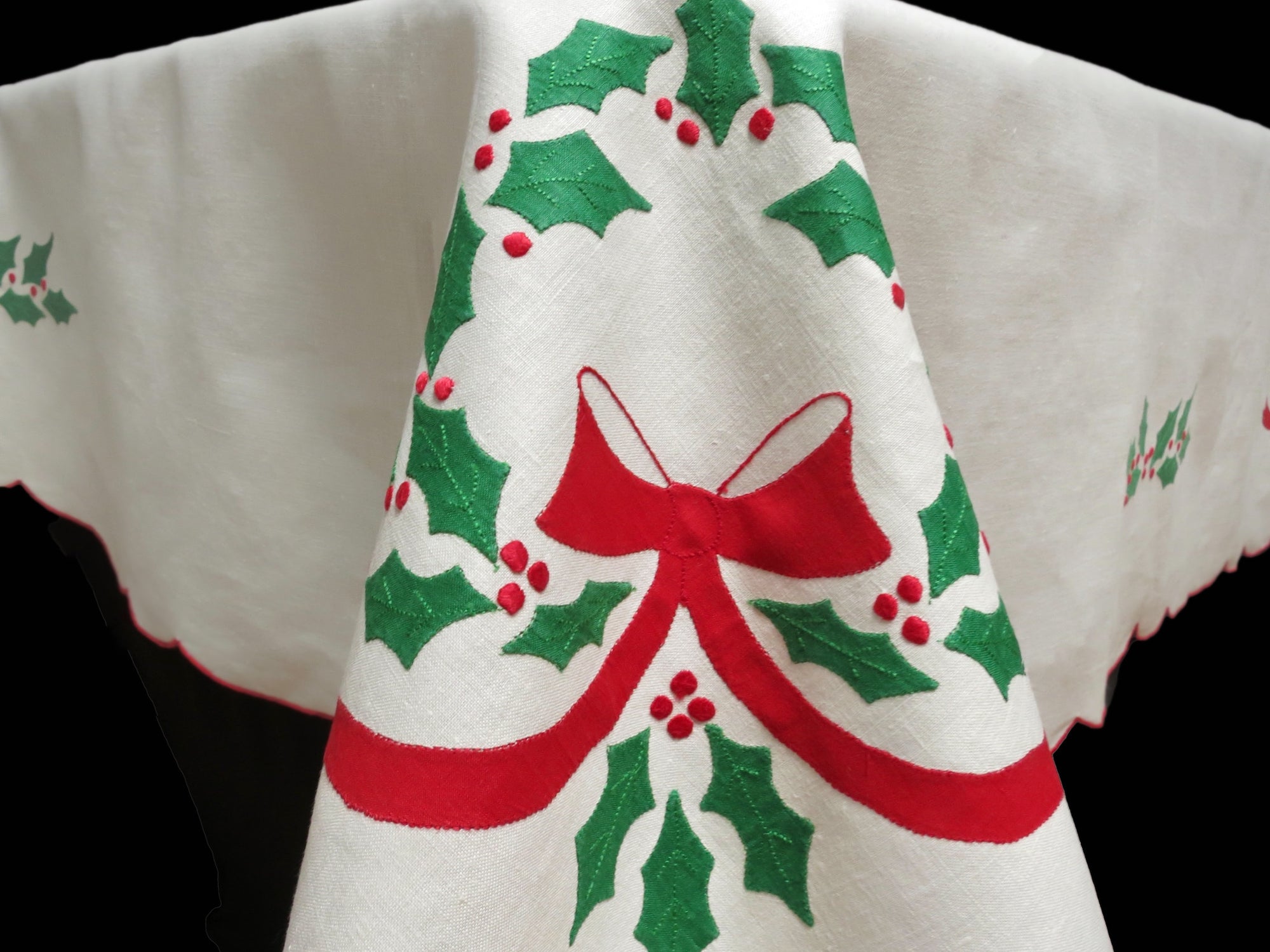 White linen tablecloth with red bow and green holly embroidery, close-up showing corner.