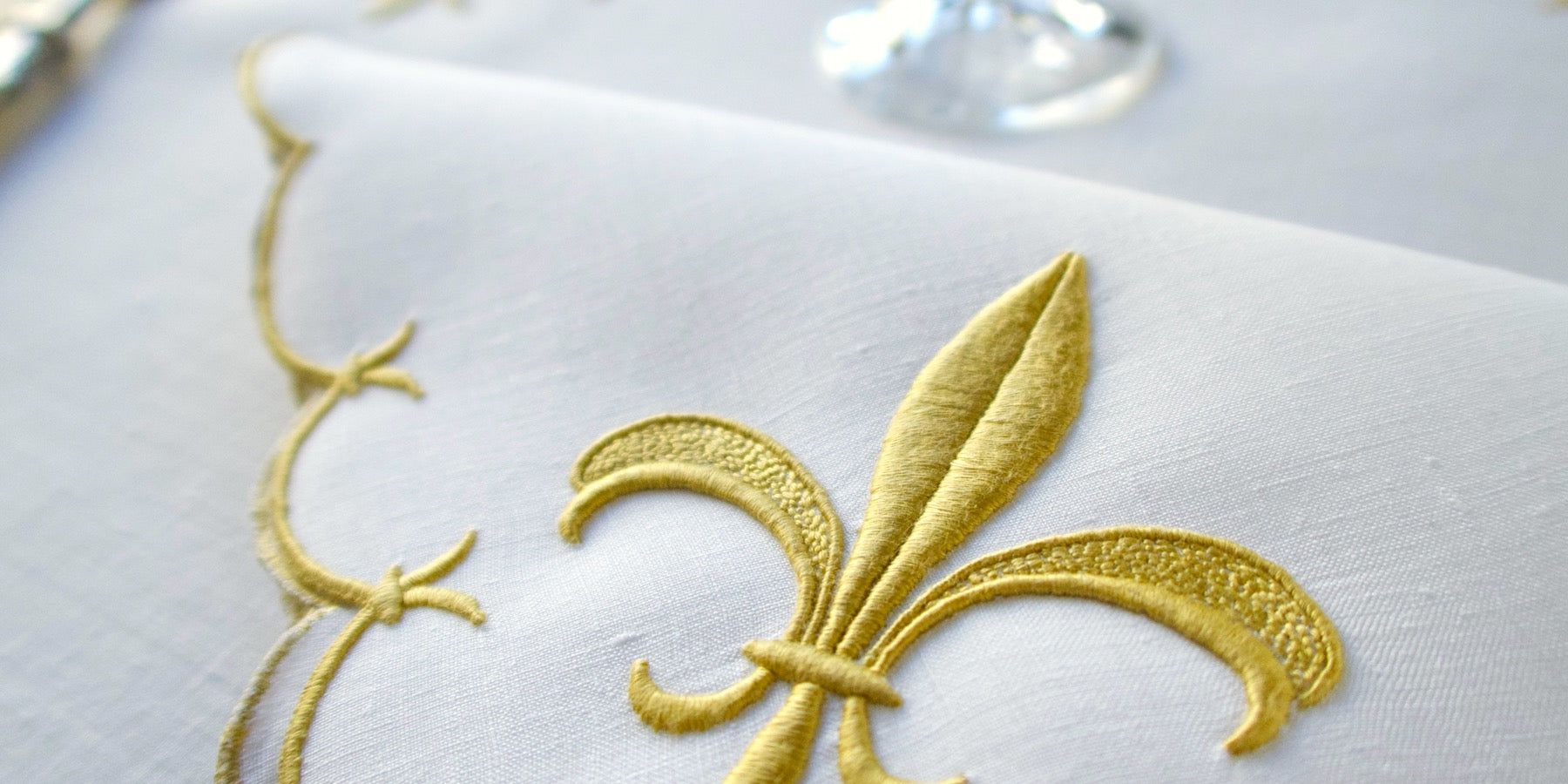 Vintage table napkin featuring Marghab's gold fleur-de-lis pattern on white linen, with a glass in the background