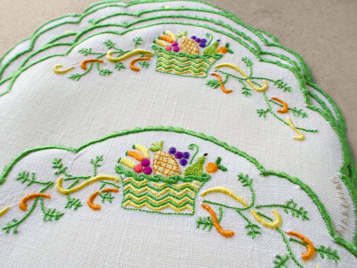 Close-up of vintage Marghab Fruit Basket cocktail round showing hand embroidered fruit basket with pineapple, grapes, and oranges on white linen with green scalloped border