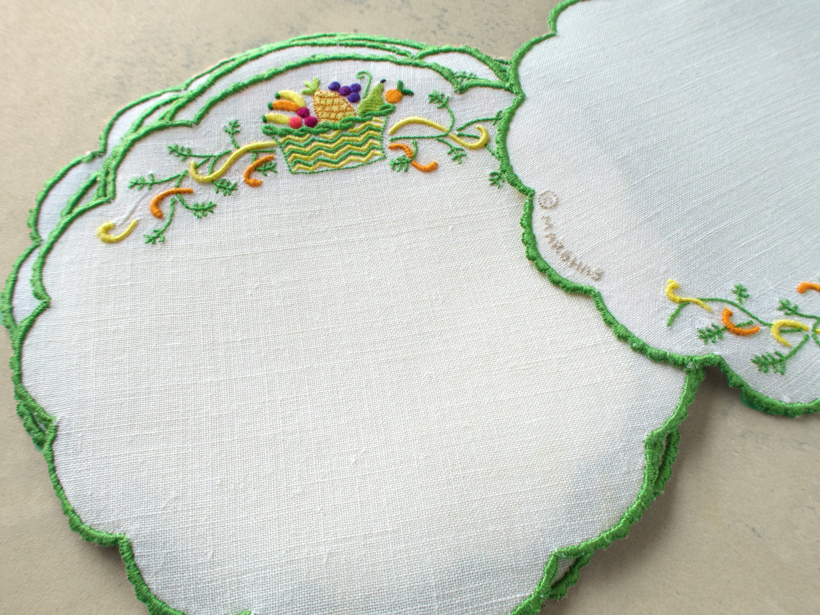 Set of vintage Marghab Fruit Basket cocktail rounds showing full round shape with hand embroidered fruit basket and green scalloped border, with the name Marghab embroidered onto the linen