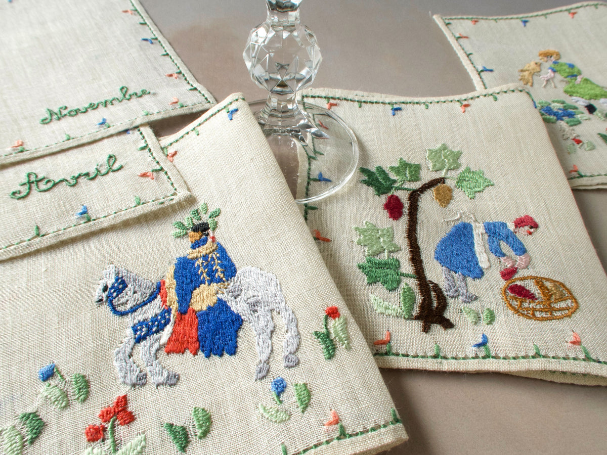 Embroidered cocktail napkins with a medieval hunting scene and a woman carrying a basket, arranged beside a clear-stemmed glass.