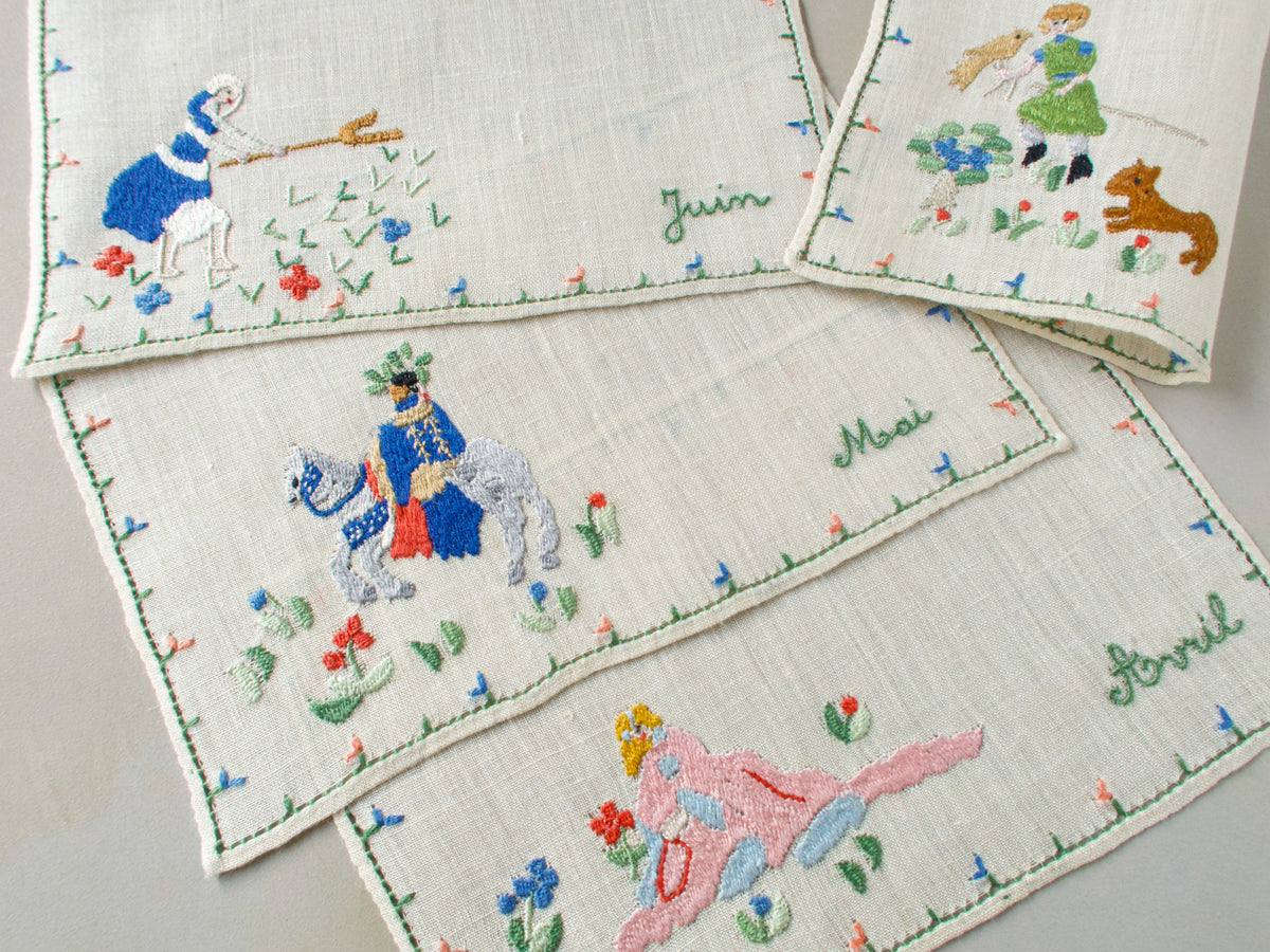 Vintage Baroness Rapisardi cocktail napkins laid flat and labeled Juin, Mai and Avril in green script, showing a haymaker, a rider on a gray horse and a reclining figure in pink beside a folded falconer napkin