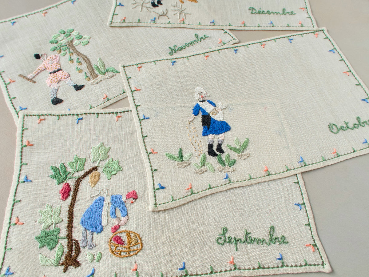 Four vintage Baroness Rapisardi cocktail napkins laid flat and labeled Septembre, Octobre, Novembre and Décembre in green script, showing a grape picker, a sower, a man knocking acorns from an oak and a hunter