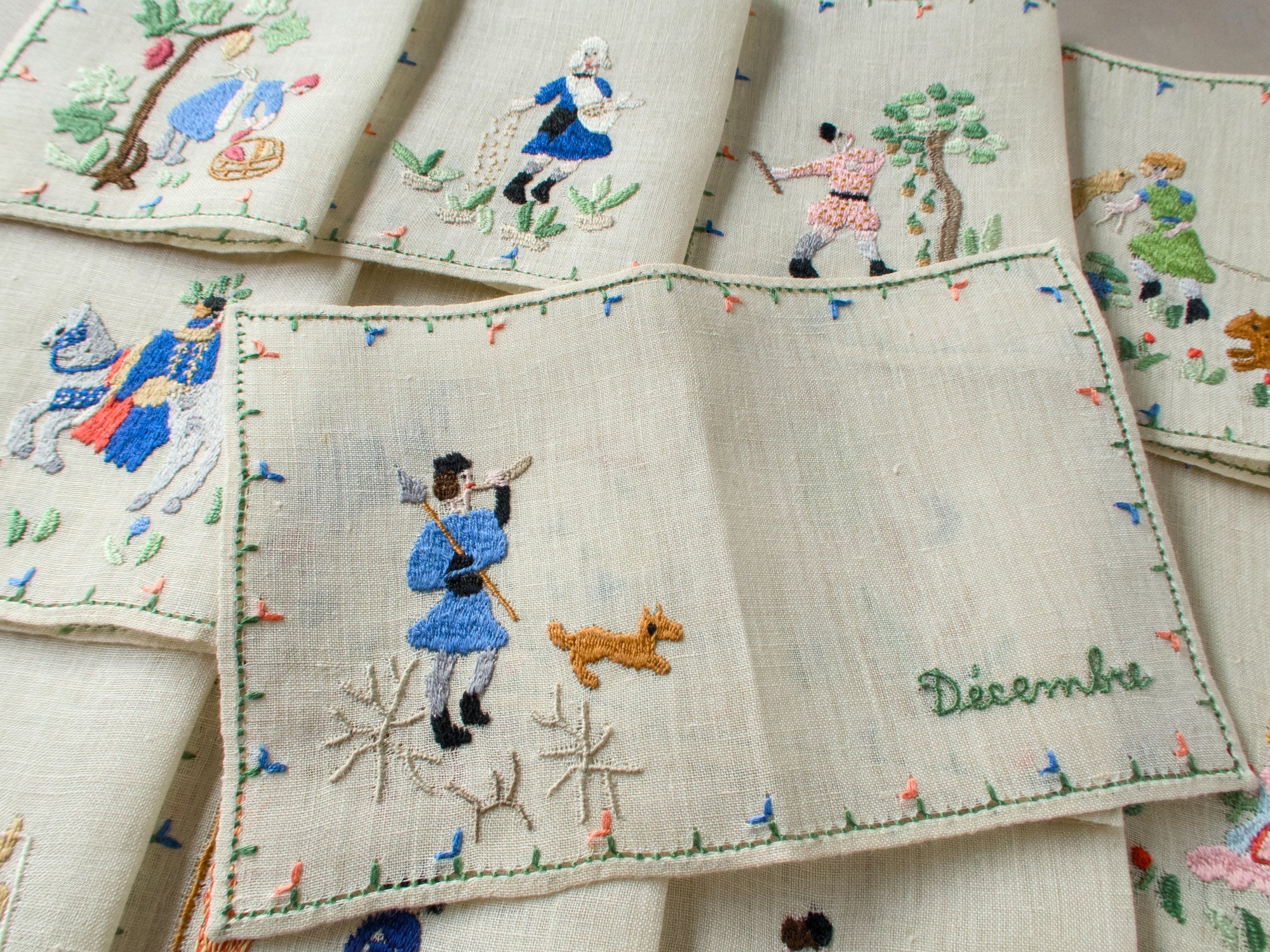 December napkin in the foreground showing a man in blue with a dog, surrounded by additional monthly scenes layered behind it.