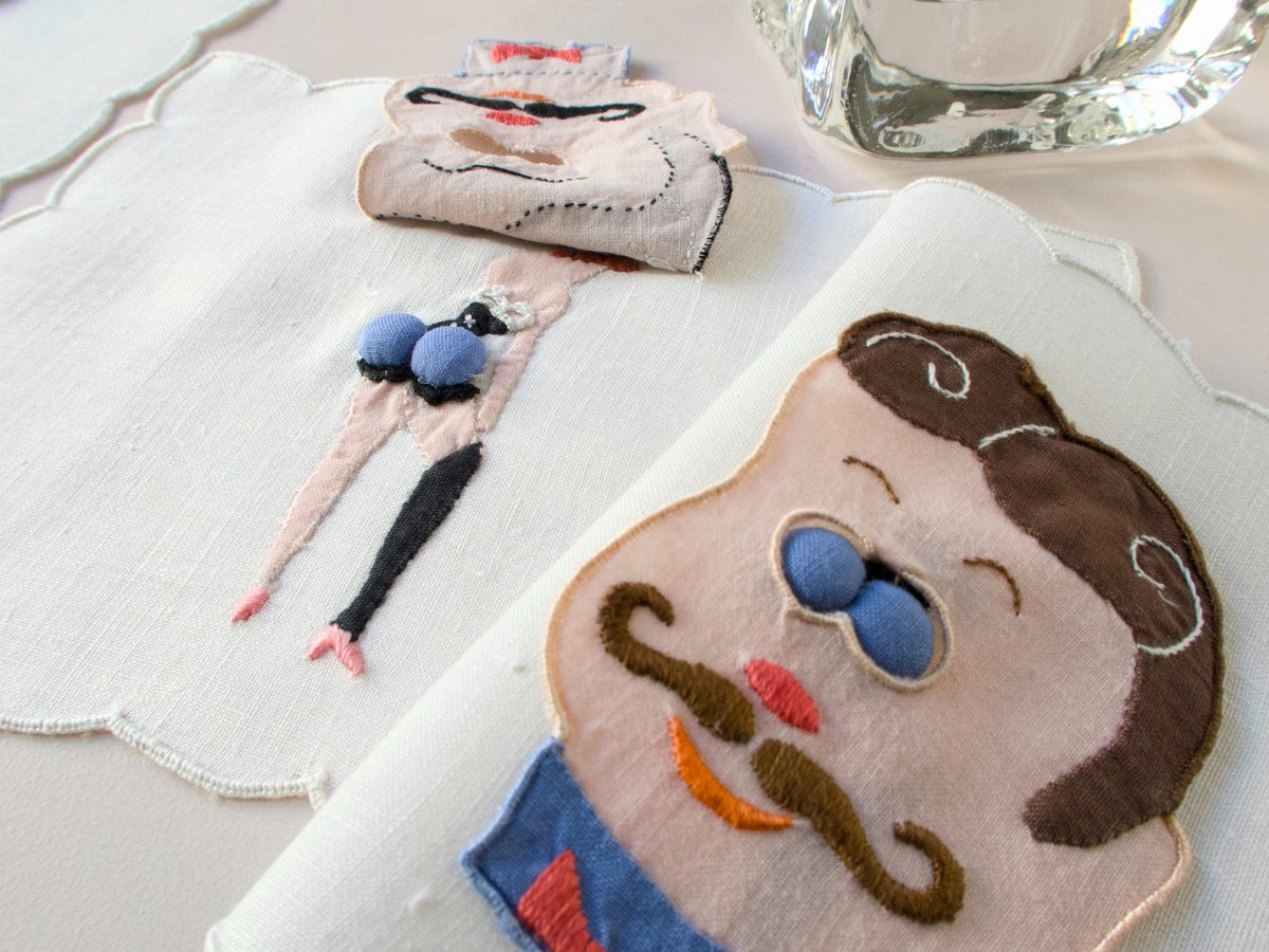 Vintage Madeira peek-a-boo cocktail napkins featuring embroidered men’s faces with blue padded ‘eyes’ that lift to reveal playful pin-up figures underneath. Close-up view showing hand embroidery and dimensional trapunto detail on linen.