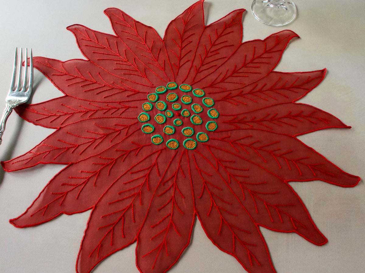 Red organdy poinsettia-shaped placemat with detailed embroidered center, photographed on a neutral background.