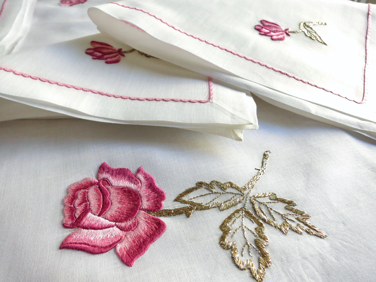 Close-up of vintage European semi-sheer cotton batiste tablecloth hand embroidered with a large pink rose and gold-accented leaves, photographed over white cloth