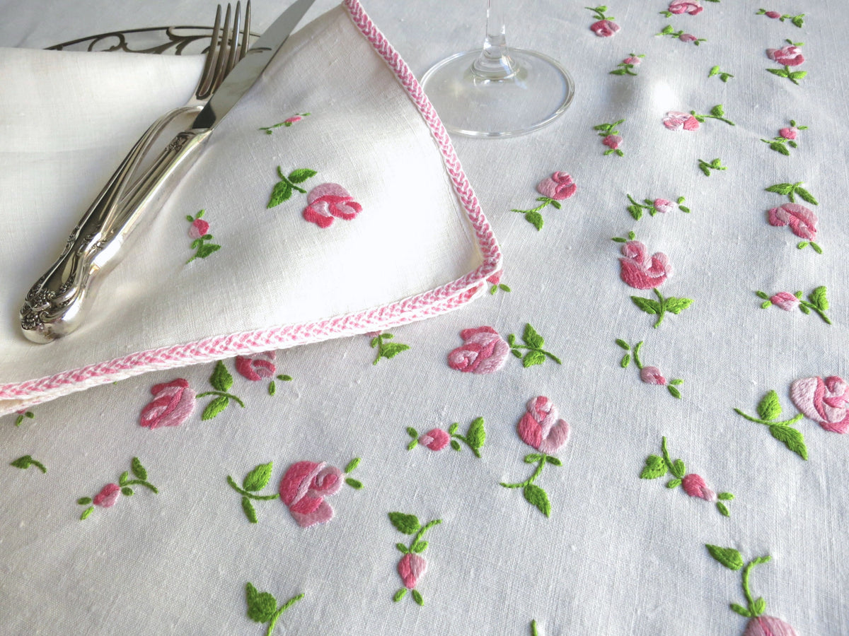 Close-up of vintage European linen napkin hand embroidered with scattered pitimini rosebuds and soft pink border, with silverware and crystal stemware