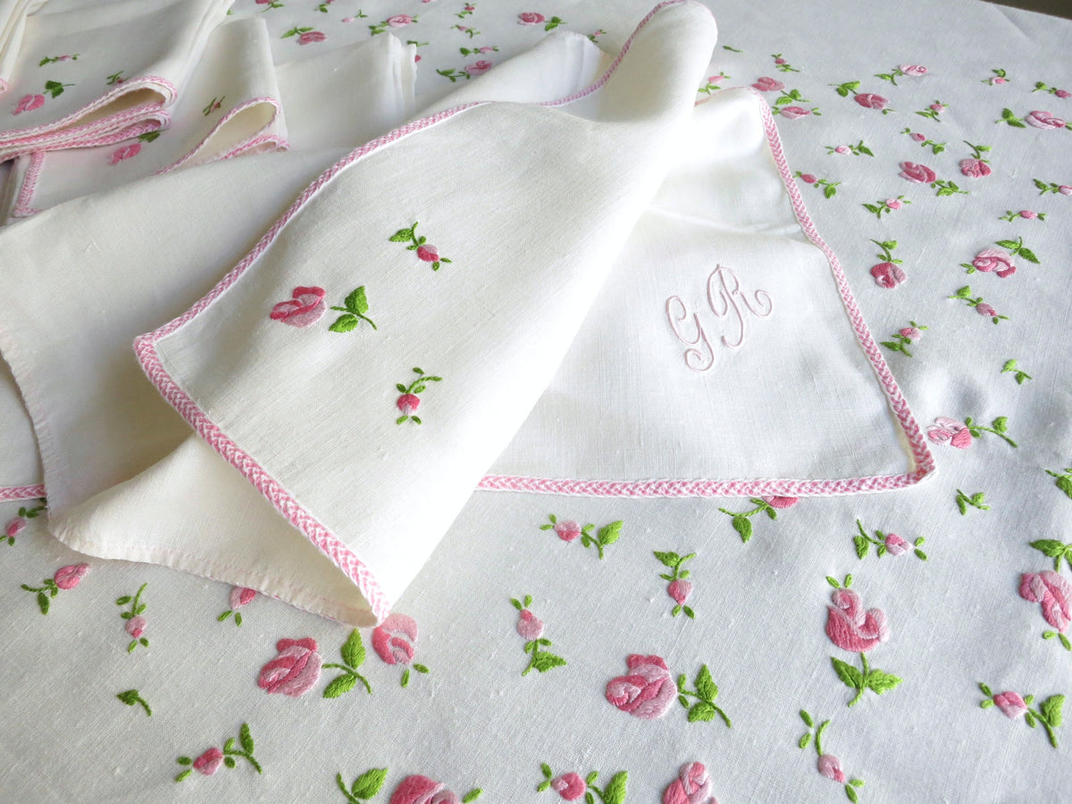 Vintage European linen napkin hand embroidered with pitimini rosebud in corner and pale pink monogram, shown folded on tablecloth