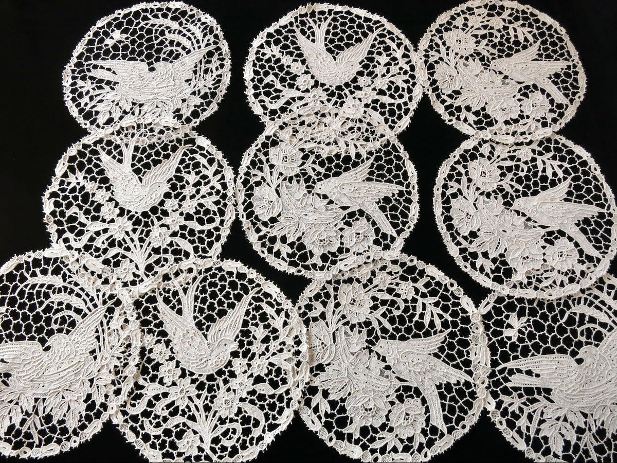 Collection of ten Italian handmade needle lace cocktail rounds, with bird scenes, laid out on a neutral background.