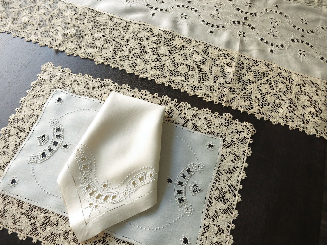 A set of vintage Milanese lace and linen placemats and napkins displayed on a table, with intricate embroidery and a white linen table runner.